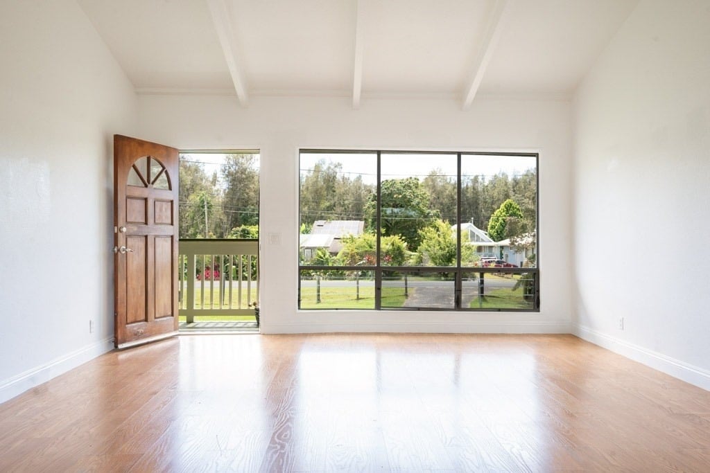 Vacant room with hardwood floors, exposed beams, and large windows overlooking a green yard.