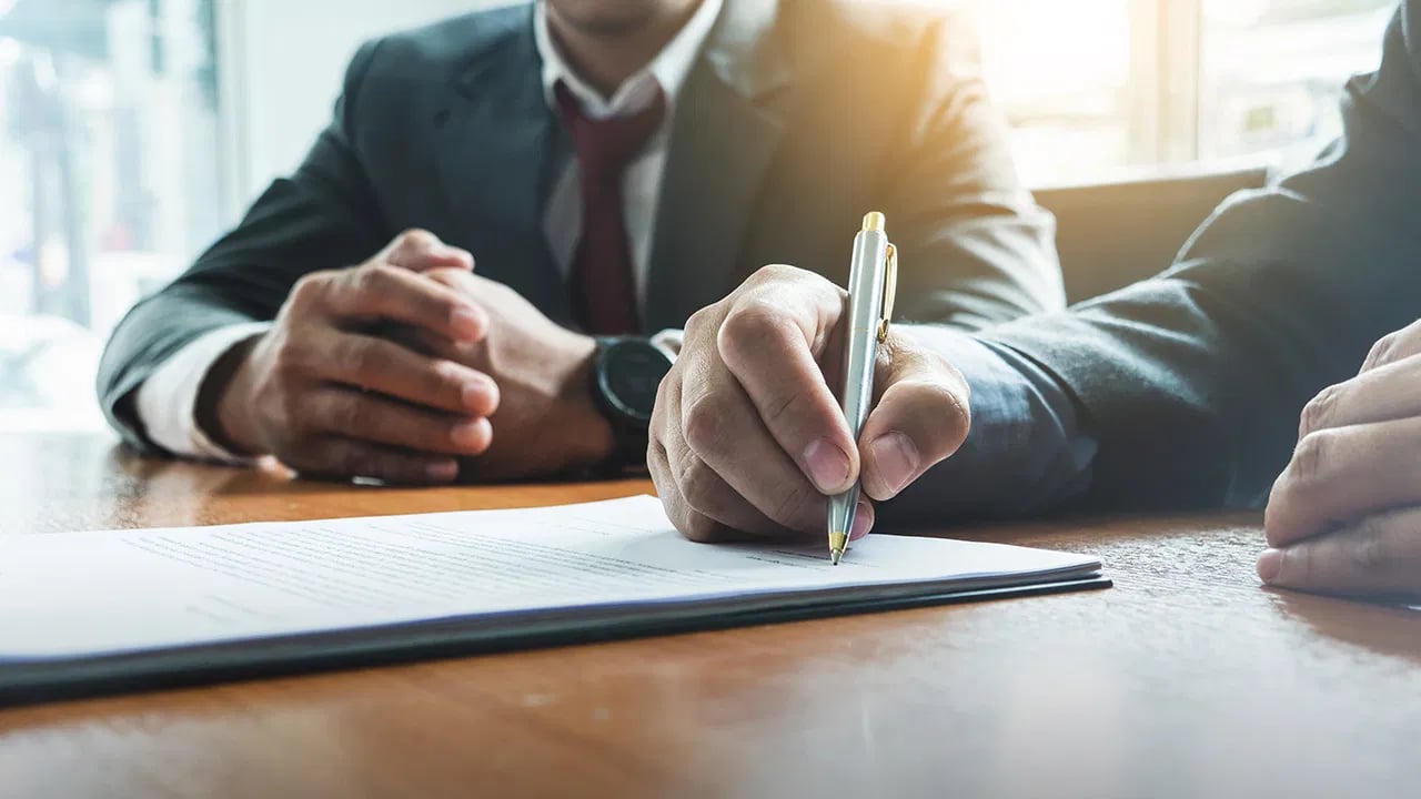A suited person signs mortgage paperwork with a silver pen at a wooden table.