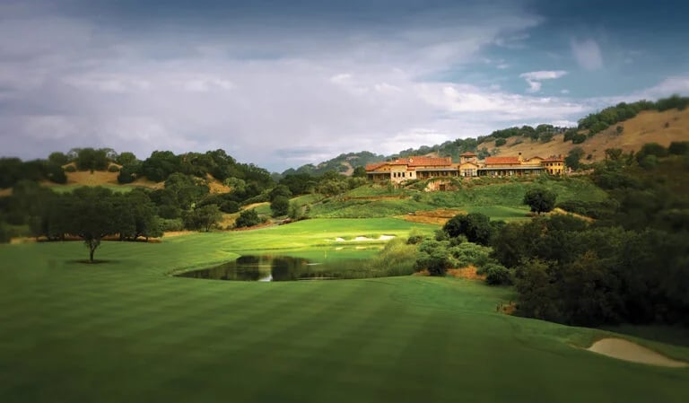 A golf course with a pond and resort buildings among green hills.