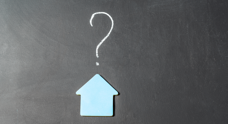 A blue house-shaped cutout sits on a blackboard. Above it, a chalk-drawn question mark suggests uncertainty or inquiry about housing or real estate.