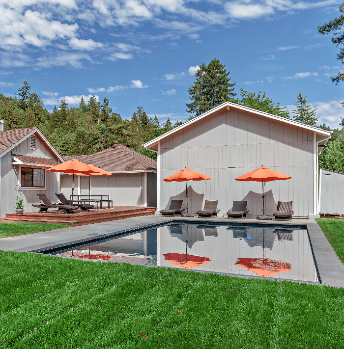 Backyard pool with orange umbrellas, lounge chairs, and wooded hills beyond.