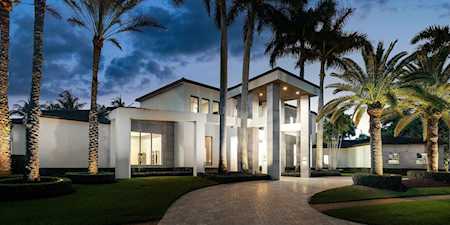 Modern luxury home with illuminated windows, a curved driveway, and palm trees at dusk.