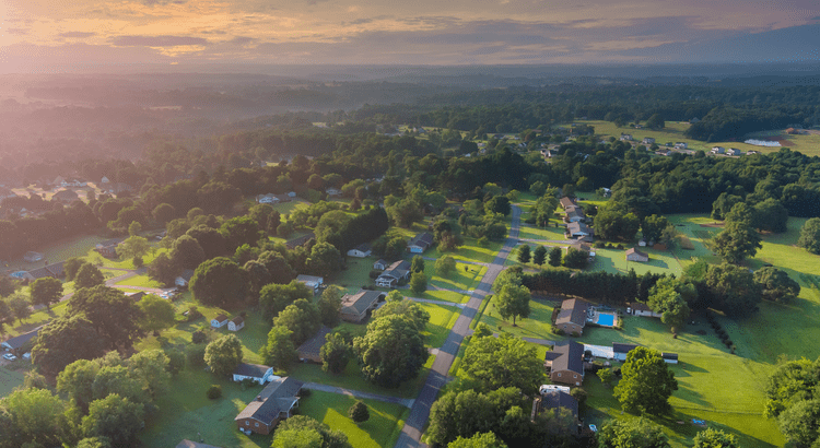 Aerial view of a serene suburban neighborhood at sunrise. Lush green trees and lawns surround houses with roads winding through. The sky is softly lit.