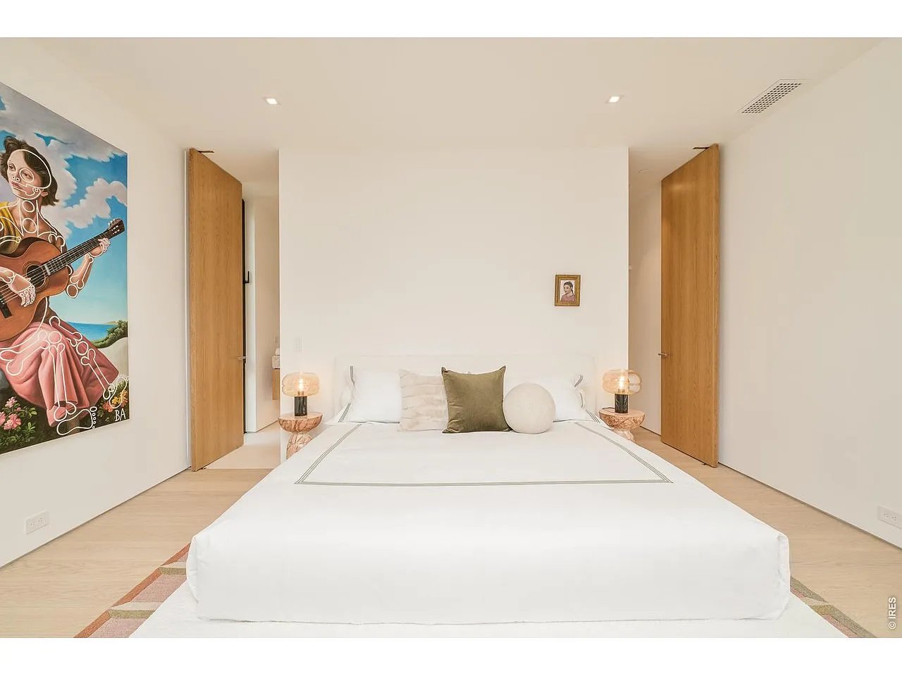 Bedroom with a large white bed, wood doors, bedside lamps, and colorful wall art.