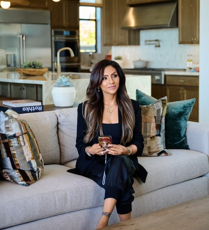 Jasmine Sunkara seated on a sofa, holding a phone in a modern kitchen.