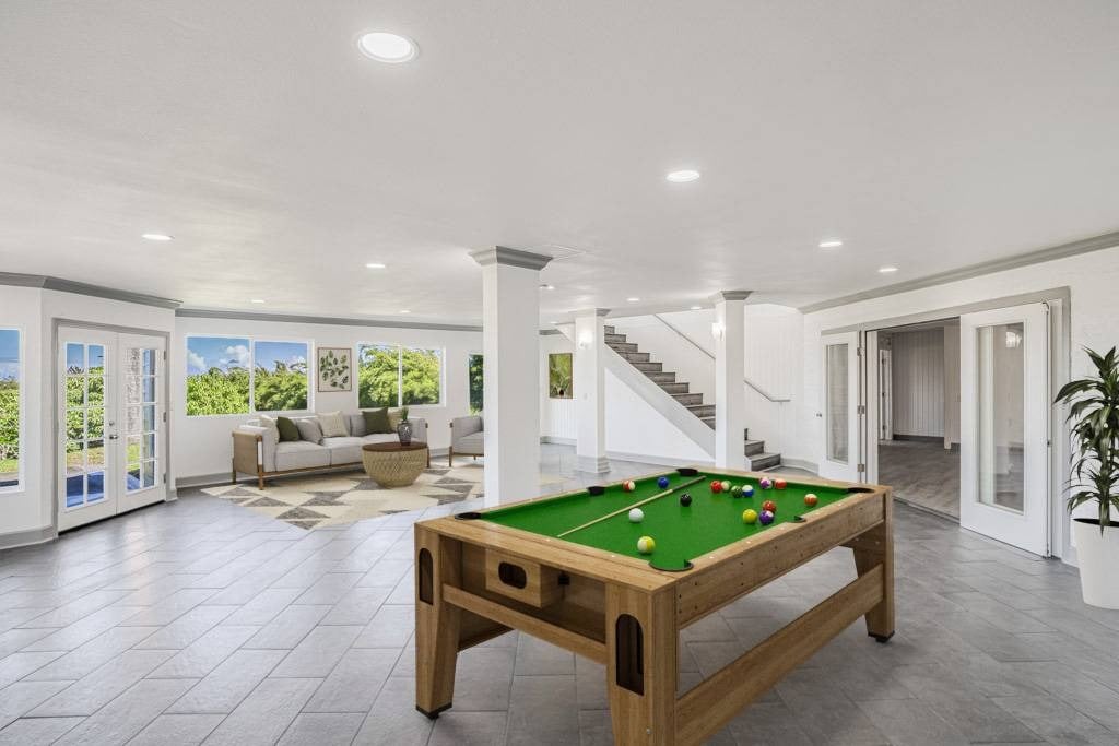 Bright recreation room with a green-felt pool table, lounge seating, large windows, and a staircase.