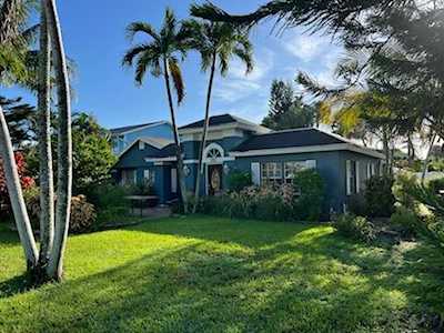 Single-story blue-gray house with a broad lawn and tall palm trees.