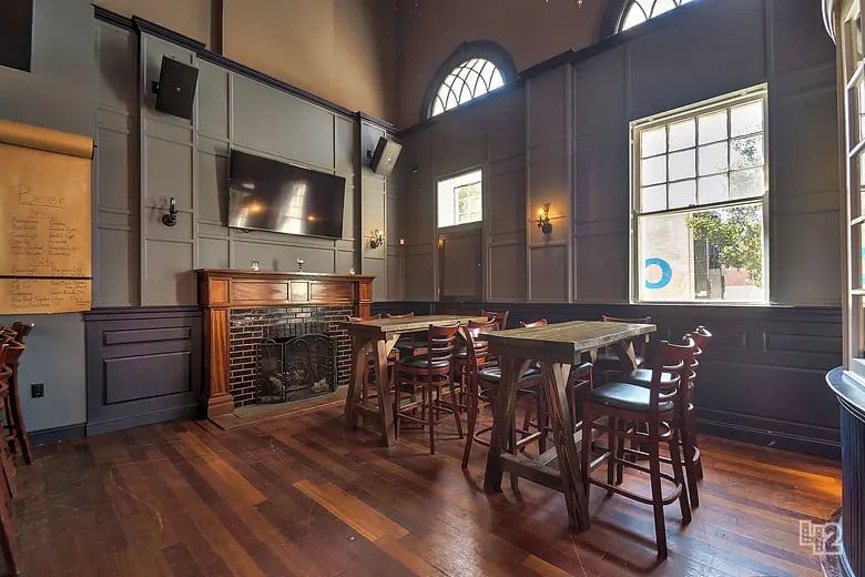 Furnished room with tall arched windows, a brick fireplace, and wooden tables and chairs.