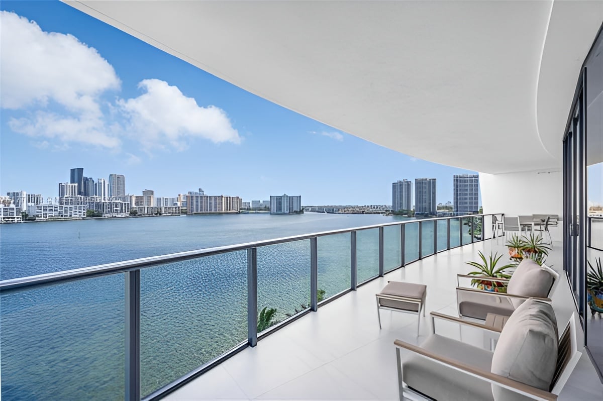 Furnished waterfront balcony with glass railing, lounge chairs, and a distant city skyline.