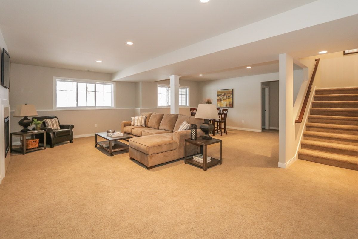 Furnished lower-level living room with a tan sectional, beige carpet, and staircase.