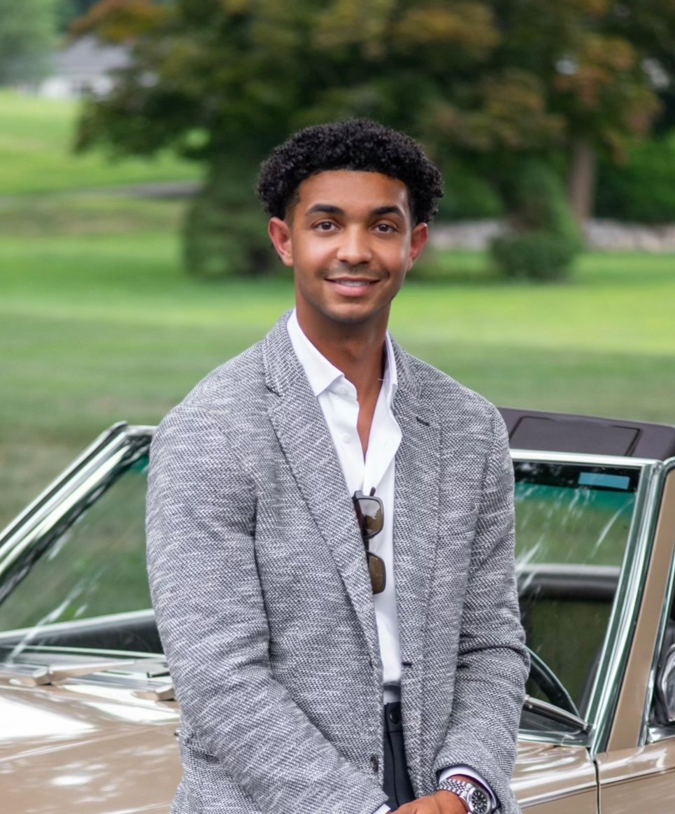 "Connecticut real estate agent in a light gray blazer standing in front of a classic convertible car outdoors."