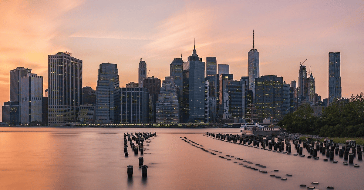 Lower Manhattan skyline across the East River at sunset, with waterfront pilings in the foreground.