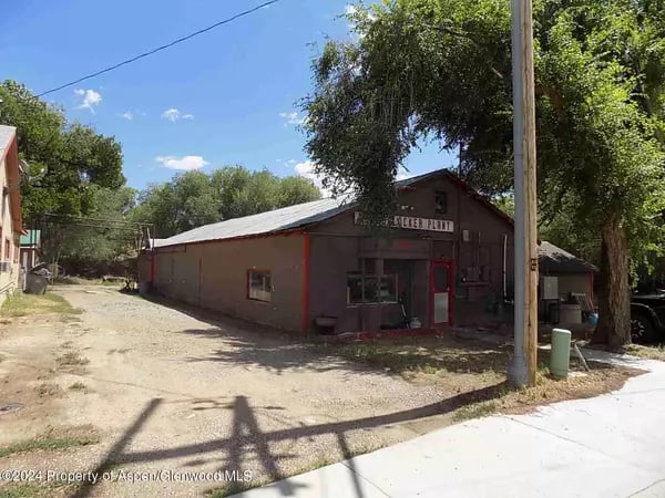 Low commercial building with red trim beside a gravel lot and leafy trees.