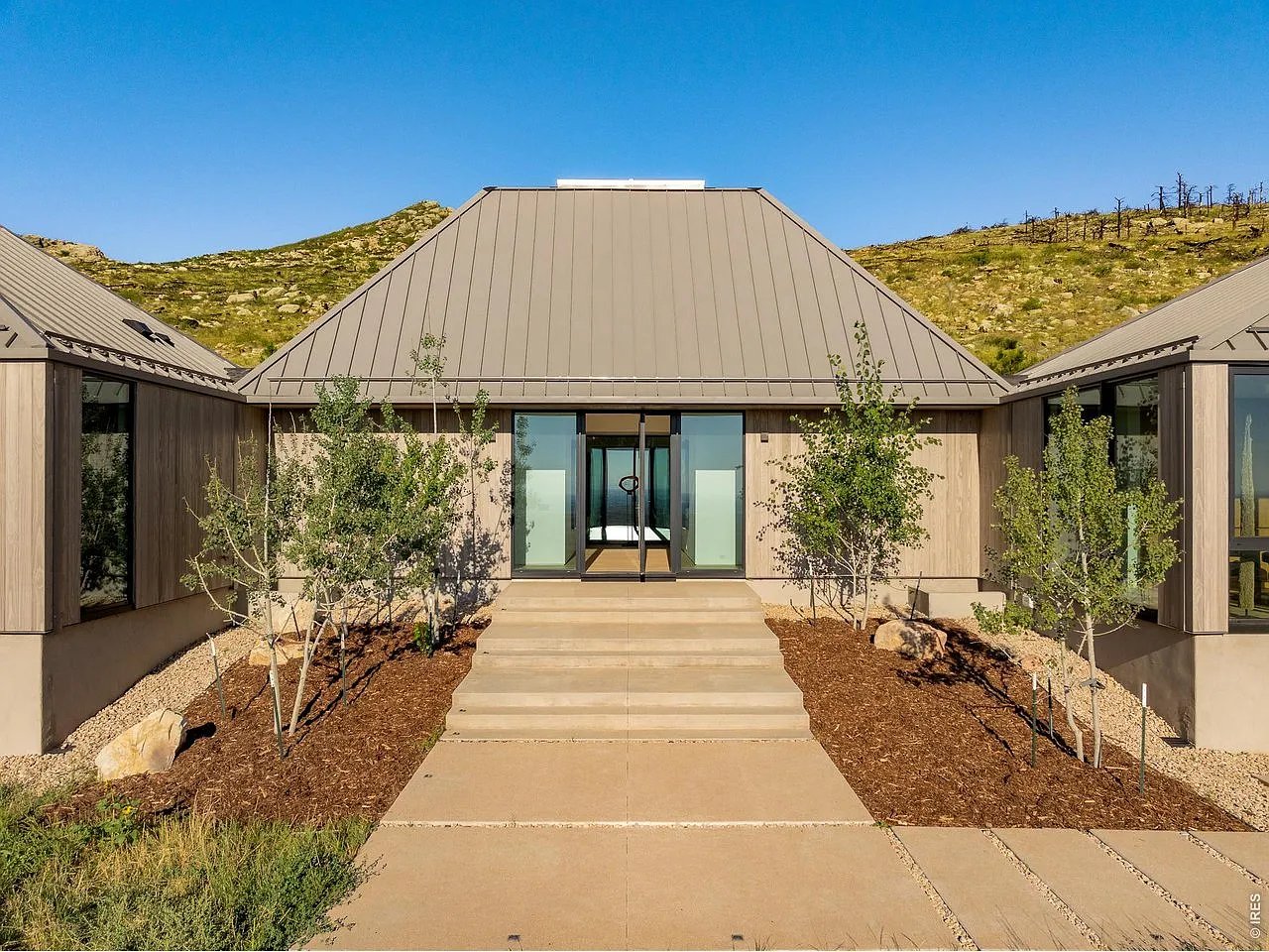 Modern home with a broad metal roof, glass entry, wide steps, and rocky hills behind it.