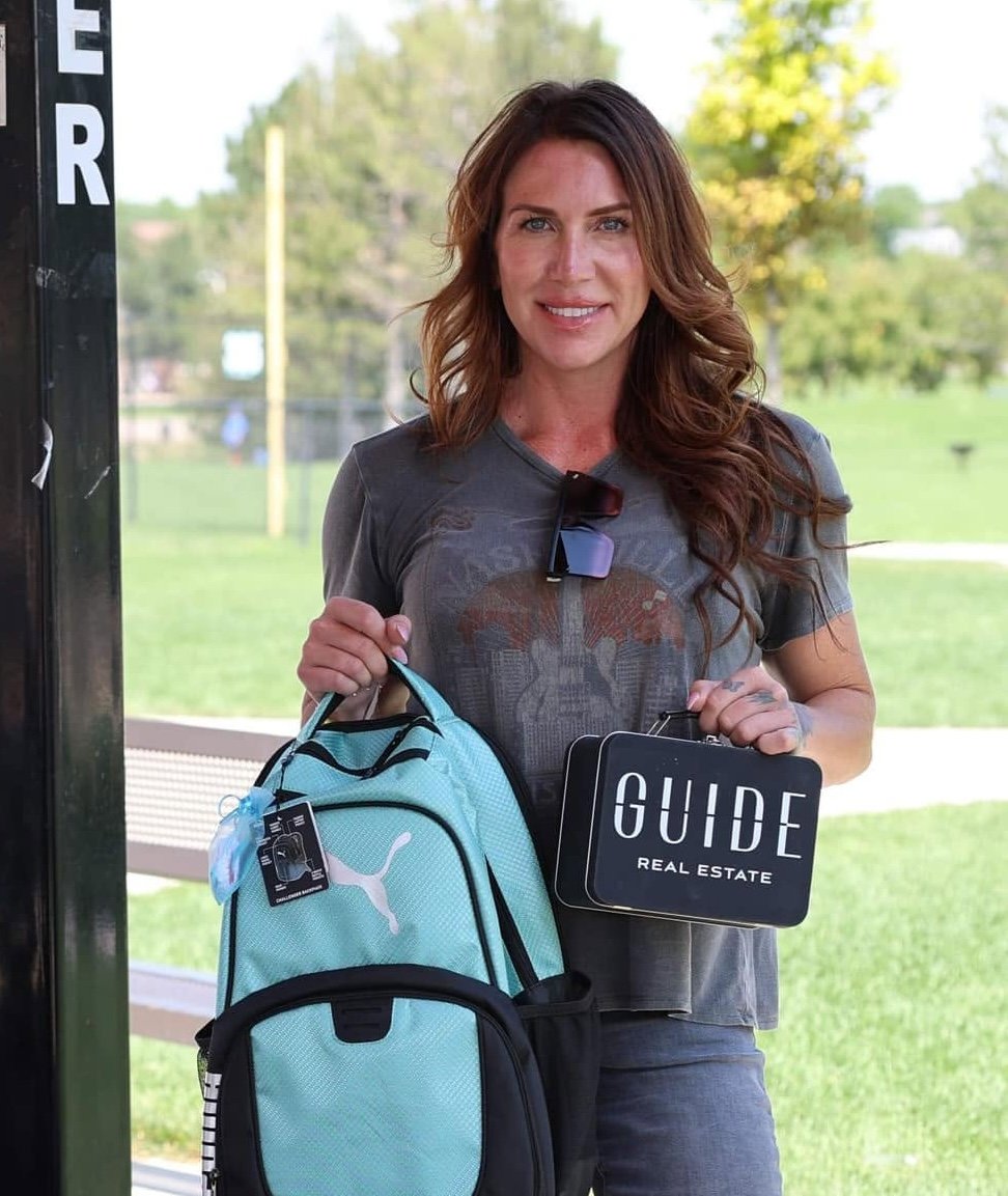 Woman holding a Guide Real Estate sign and turquoise backpack outdoors.