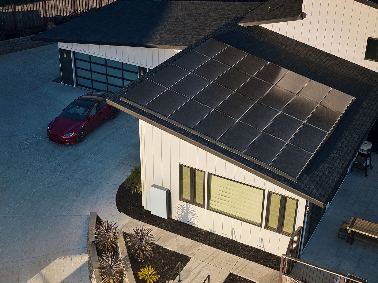 Aerial view of a white board-and-batten house with a low-slope roof covered in black solar panels, a red car in the driveway, and a white Tesla Powerwall mounted on the exterior wall beside a row of windows.