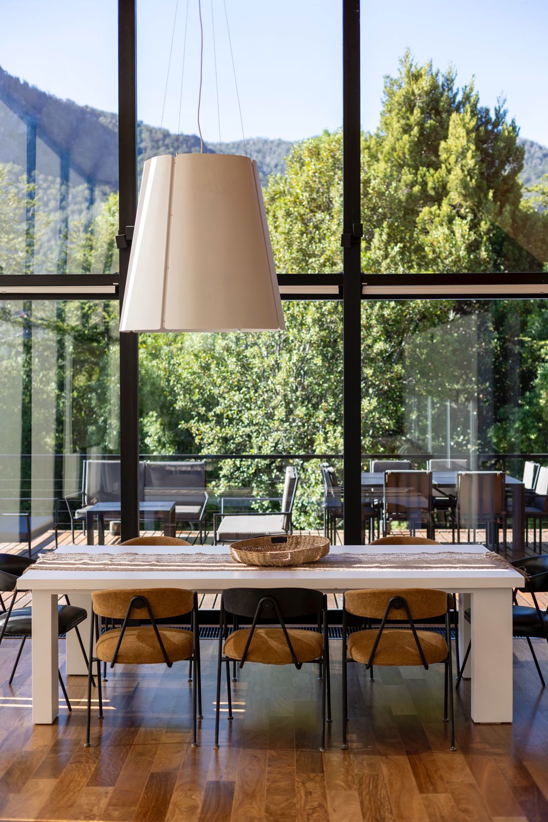 Dining room with a white table and pendant light beside floor-to-ceiling windows overlooking wooded hills.