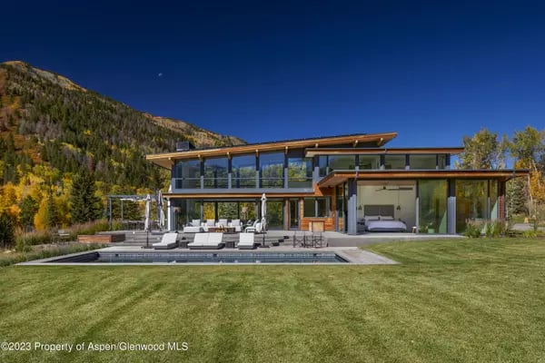 Modern glass-and-wood home with a swimming pool, framed by autumn foliage and a mountain slope.