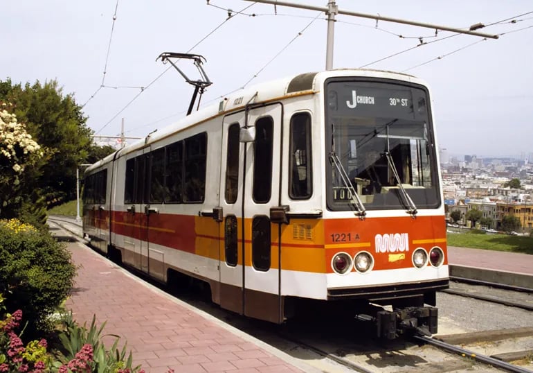 A Muni J Church streetcar passing by Mission Dolores Park with people walking towards the park entrance