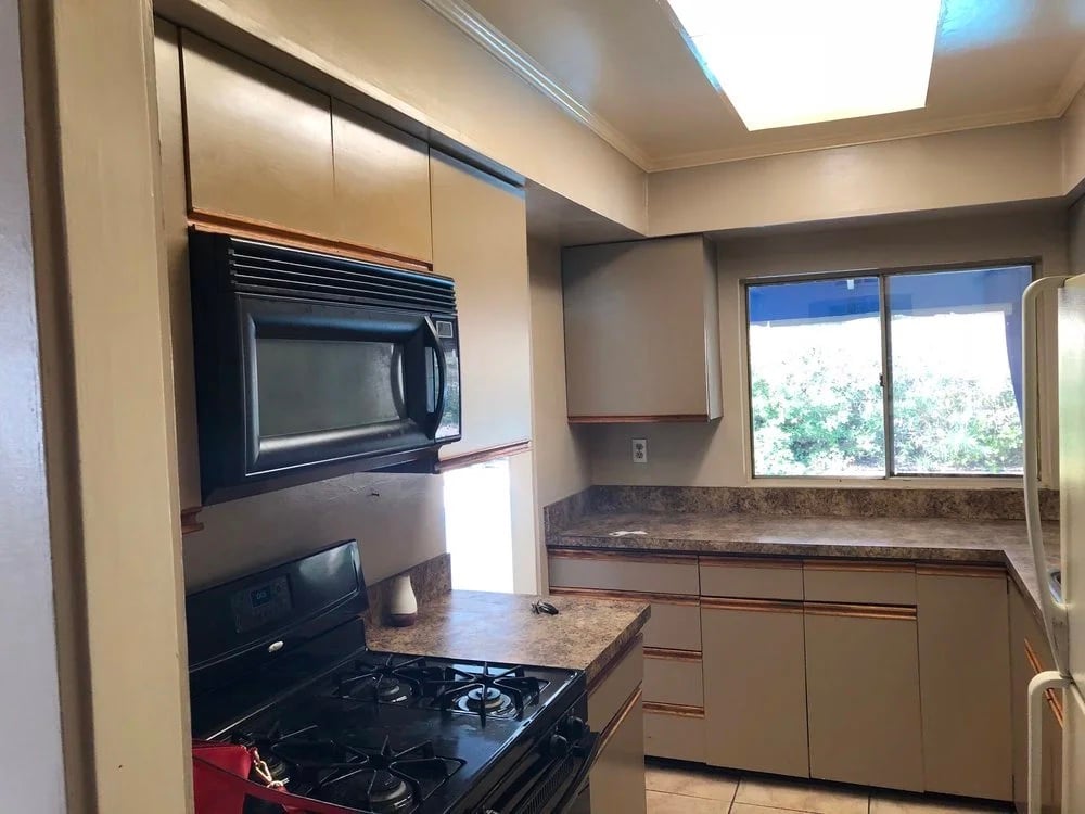 The same Palm Springs kitchen before the remodel: beige cabinets with wood trim, a black over-range microwave, laminate counters, and a fluorescent ceiling panel.