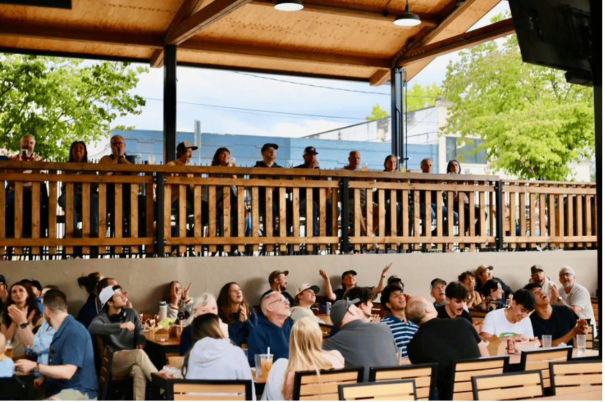 Crowded patio with seated guests and people standing along an upper wooden railing.
