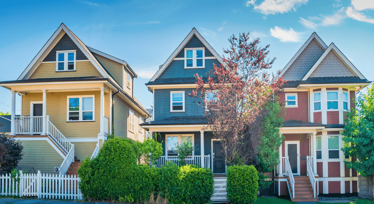 Three colorful Victorian-style houses with steep roofs, front porches, and a picket fence under a bright blue sky. Green shrubs line the walkway.