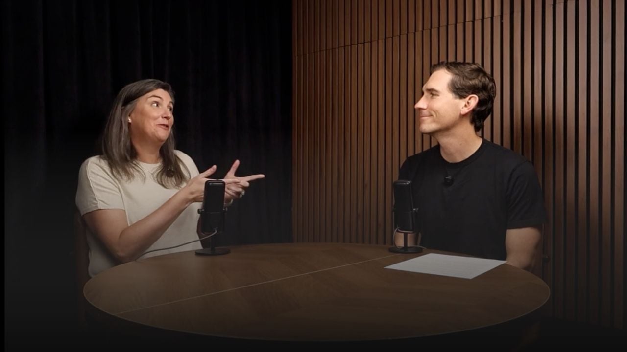 Two podcast hosts talk across a round wooden table with microphones.