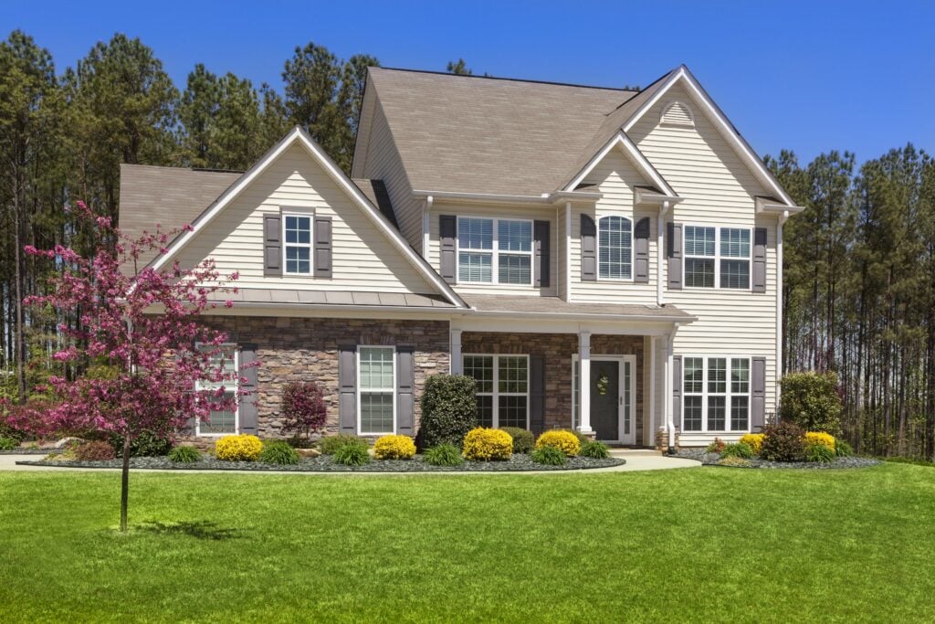 Two-story cream-sided house with stone accents, gray shutters, and a landscaped lawn.