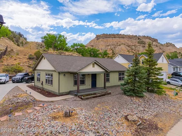 Single-story green home with a covered porch, rocky yard, and mesa behind it.