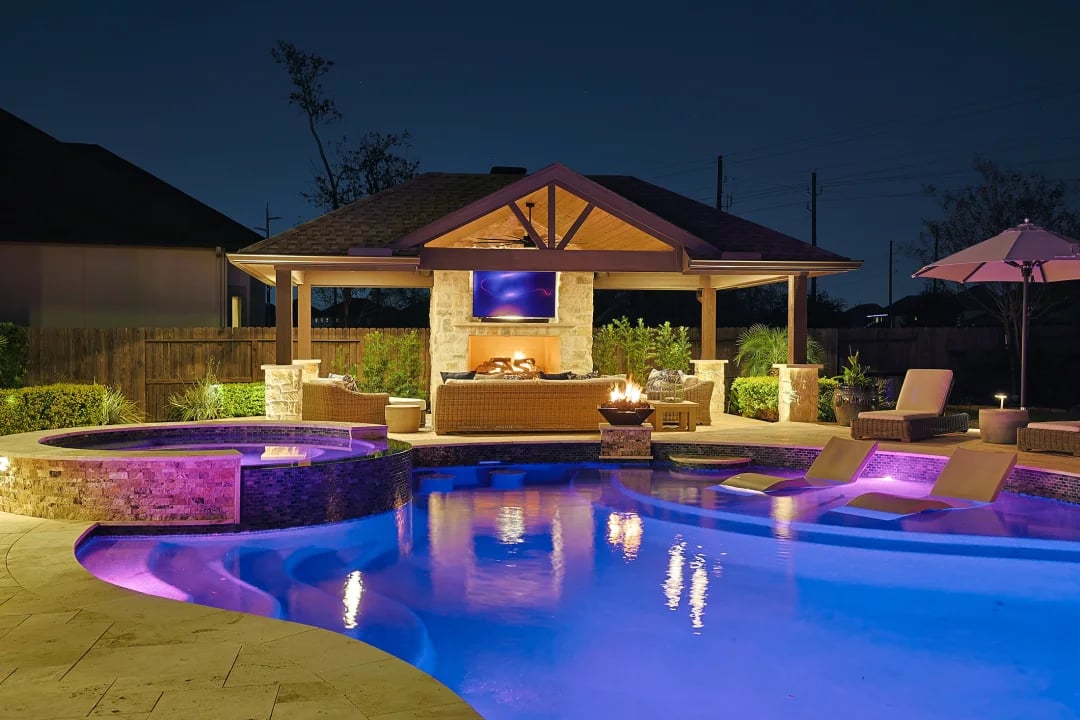 Illuminated backyard pool with tanning ledges and a covered stone pavilion at night.