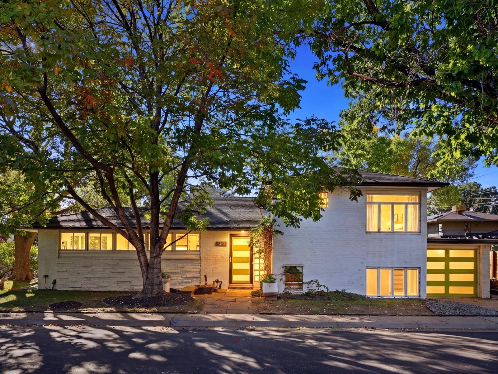 Mid-century modern home with a large front-yard tree, glowing windows, and an attached garage at dusk.