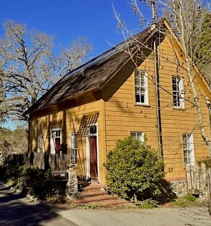 Yellow two-story house with a steep shingle roof, stone foundation, and red front doors.