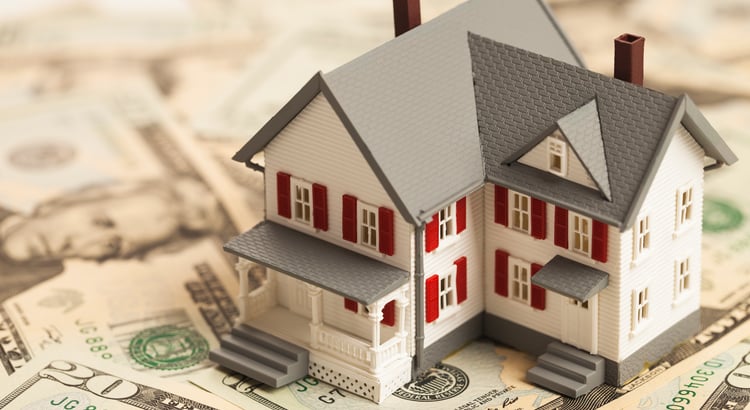 Miniature white house with red shutters standing on U.S. dollar bills.