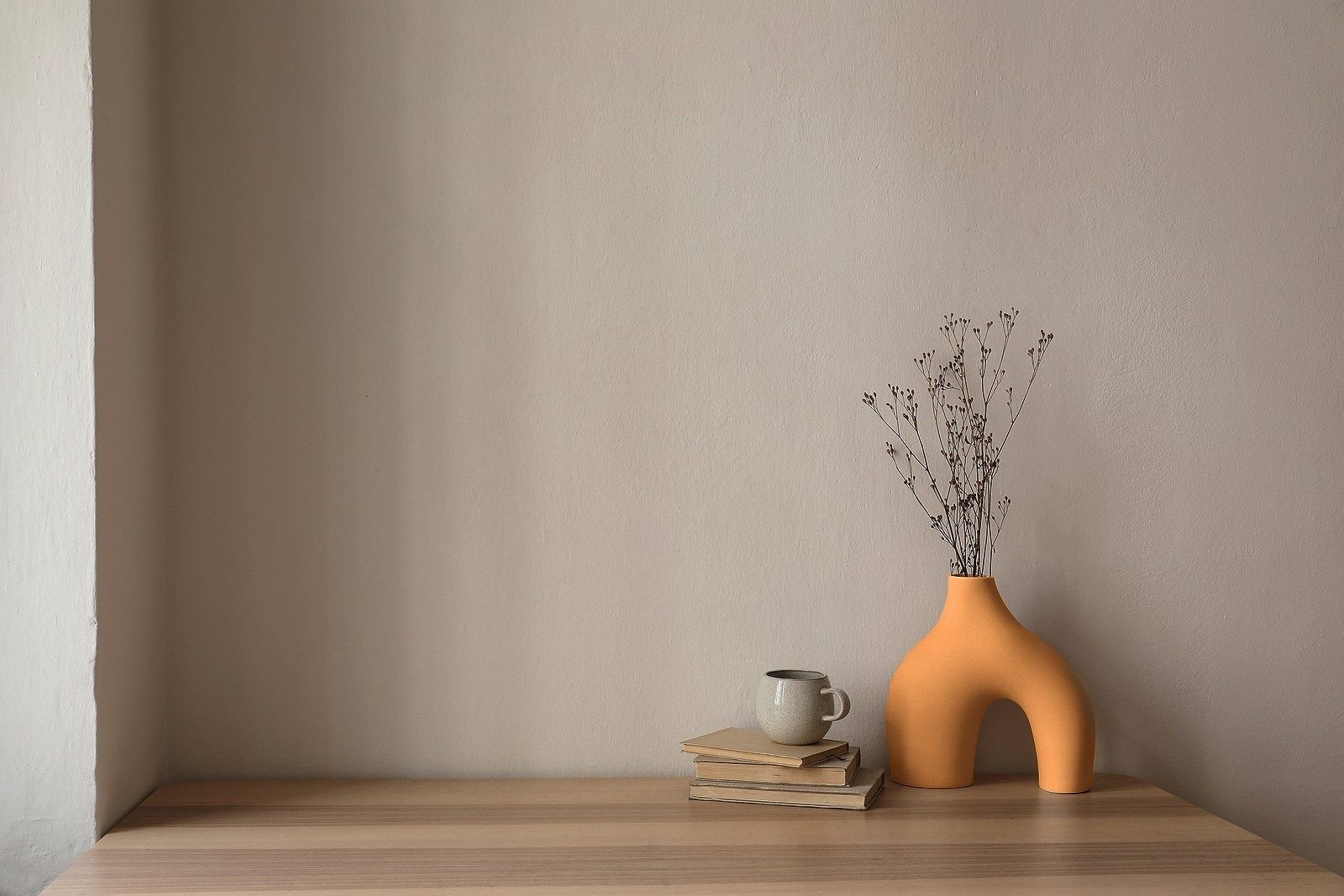 Console table with an orange sculptural vase, dried stems, books, and a ceramic mug against a pale wall.