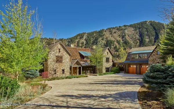 Stone mountain home with a curved driveway, mature landscaping, and forested mountain backdrop.
