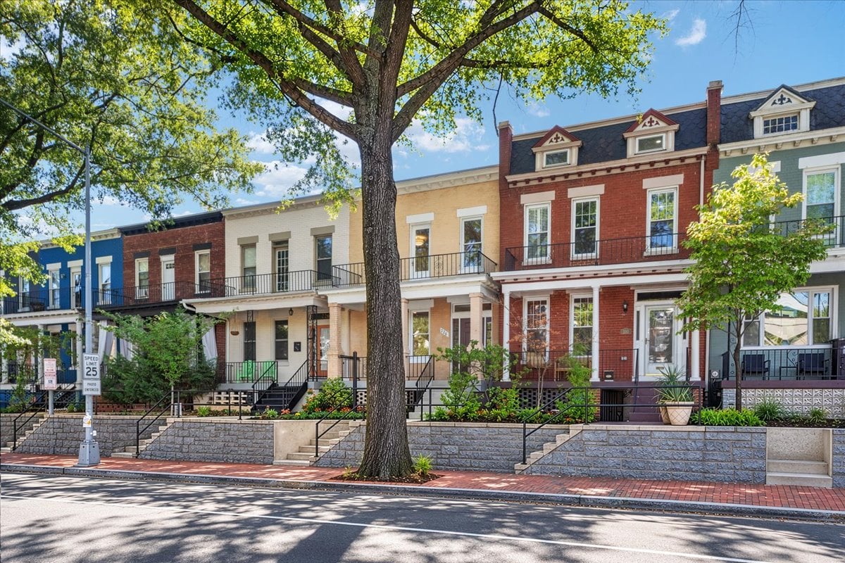 Attached townhouses with brick façades and front stoops, partly shaded by a large street tree.
