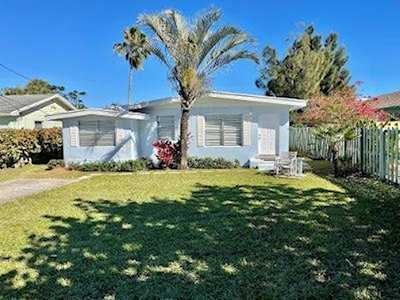 Single-story house with shuttered windows, a broad lawn, palm tree, and patio chairs.