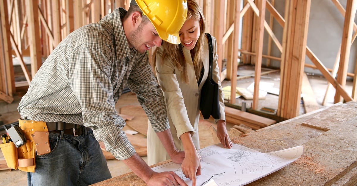 A woman in a business suit and a contractor in a hard hat point together at blueprints on a table inside an unfinished home.