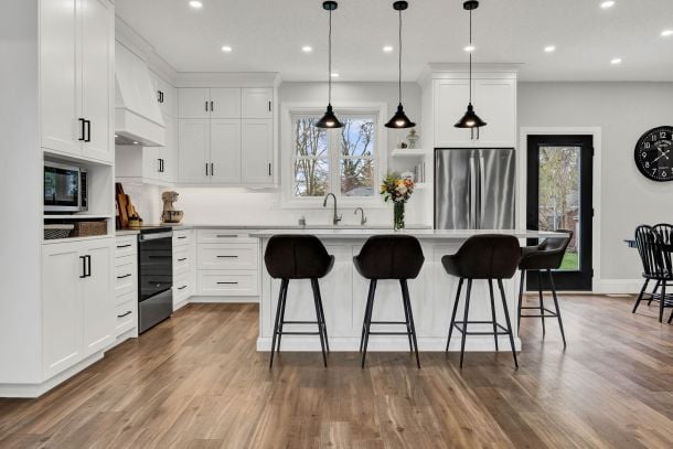 Bright white kitchen with a large island, three dark stools, pendant lights, and wood-look flooring.