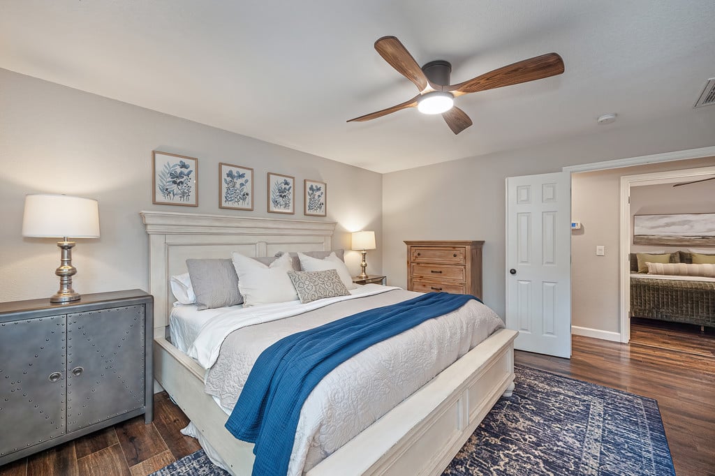 Staged bedroom with a large white bed, blue throw, patterned rug, and wood ceiling fan.