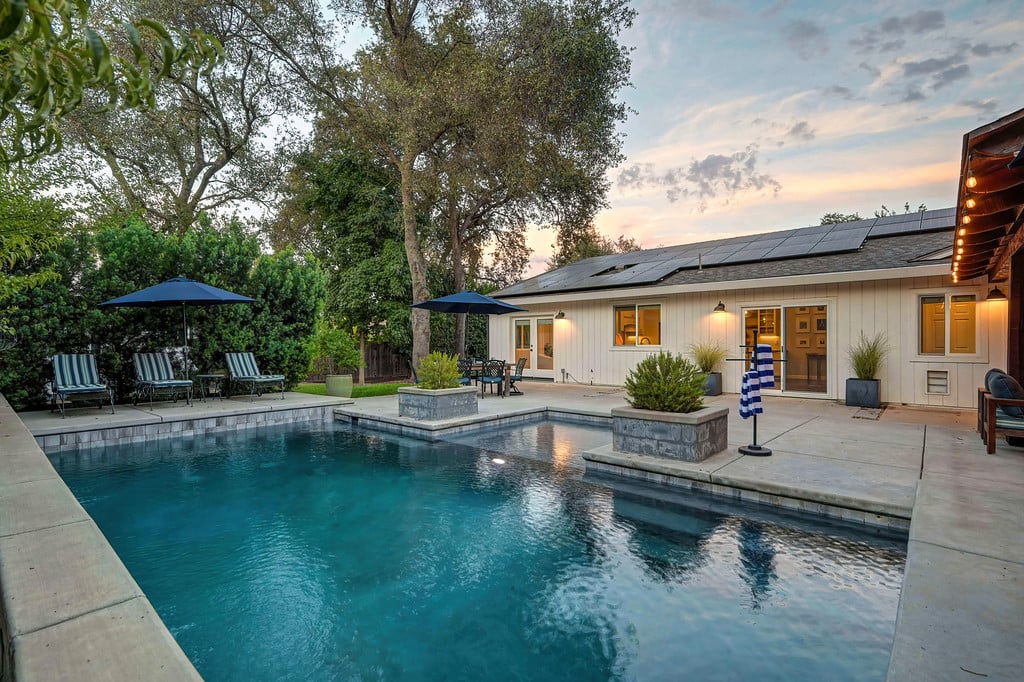 Backyard pool beside a single-story home with solar panels and patio seating at dusk.