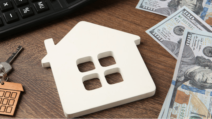 White house-shaped model beside a keyboard, keys, and hundred-dollar bills on a wooden desk.
