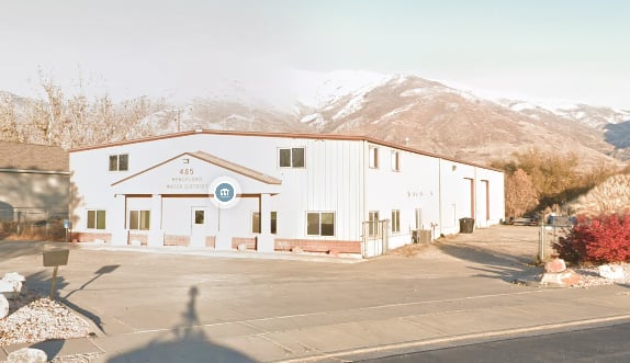 Long white commercial building with a paved parking area and snow-dusted mountains behind it.