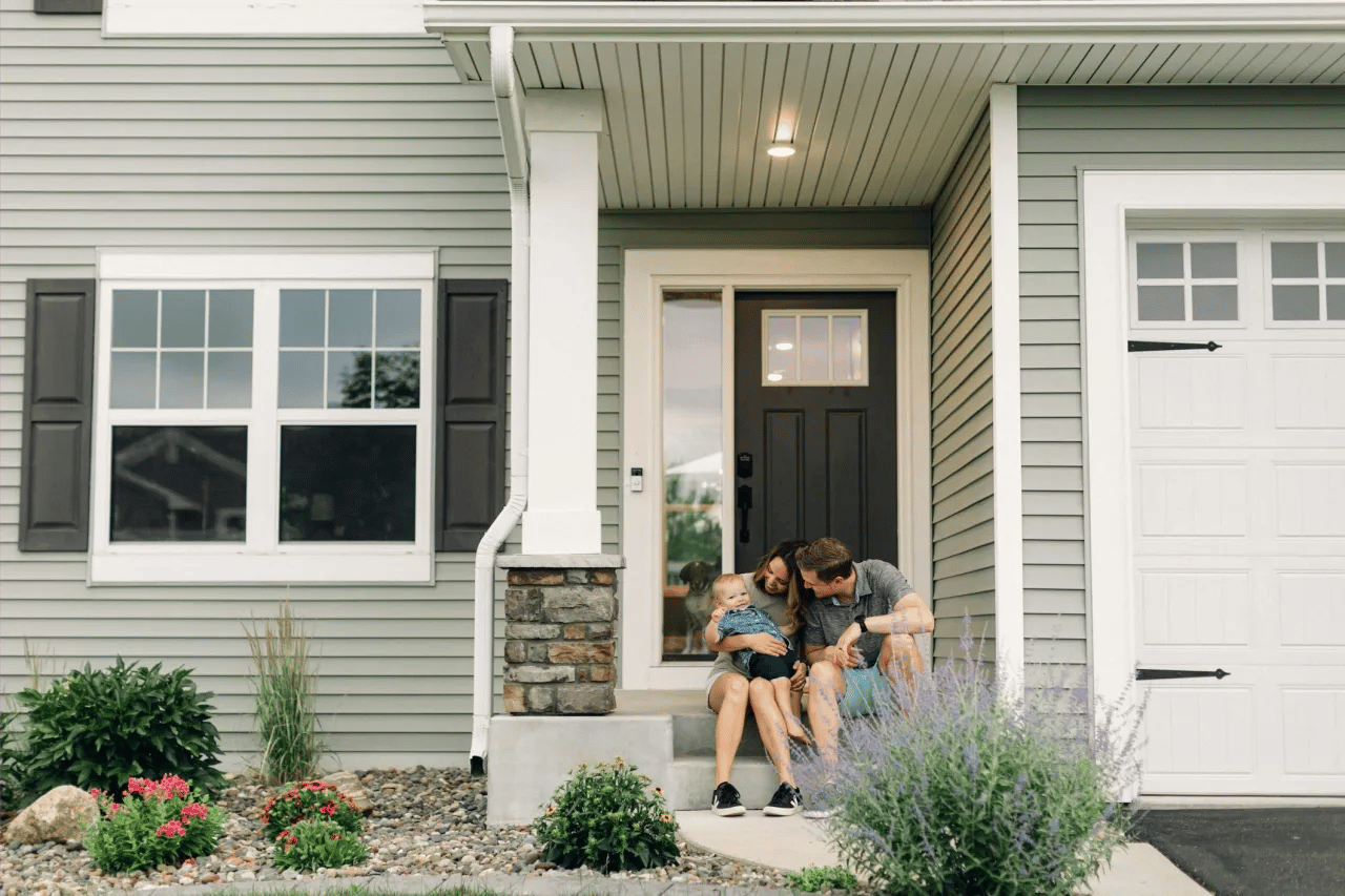 Gray-sided house with a family and baby on the front step beside landscaped flower beds.