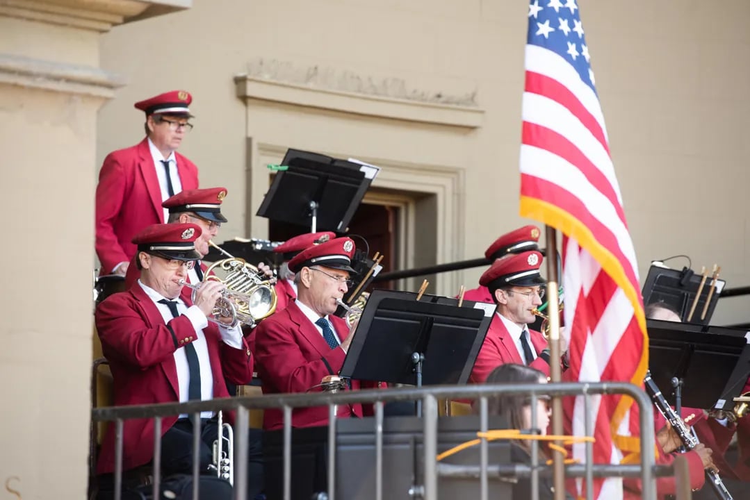 Members of the Golden Gate Park Band in red uniforms playing brass and woodwind instruments next to an American flag during a live outdoor concert in San Francisco.