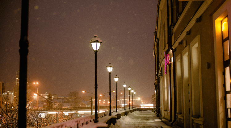 Snowy sidewalk lined with glowing street lamps beside a building at night.