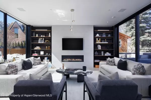 Furnished living room with a linear fireplace, built-in shelves, and large windows overlooking snow-dusted trees.