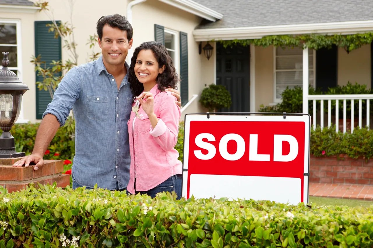 A house with a red SOLD sign and a smiling couple holding keys.