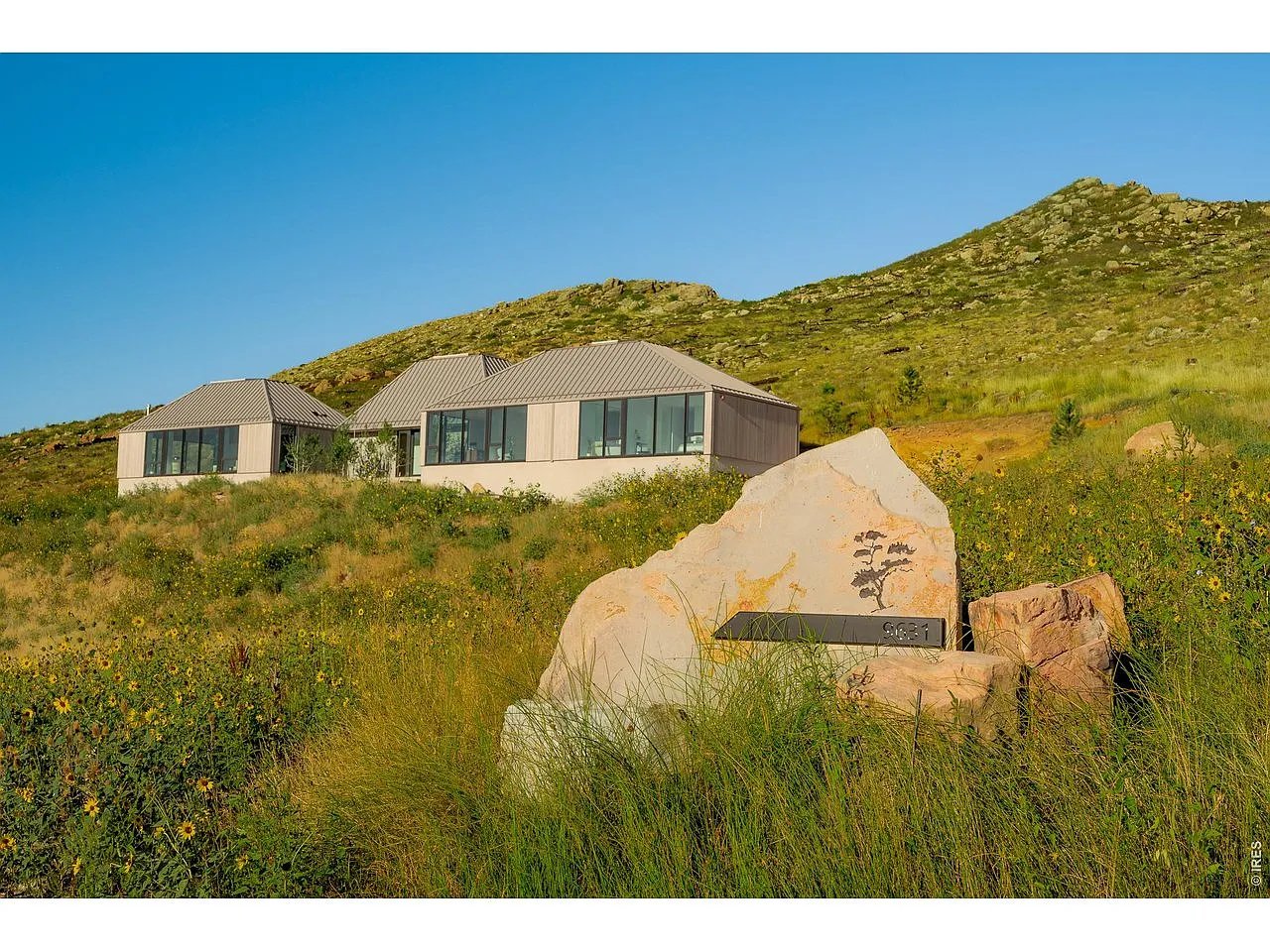 Modern home on a grassy hillside, with a large stone address marker and rocky ridge.