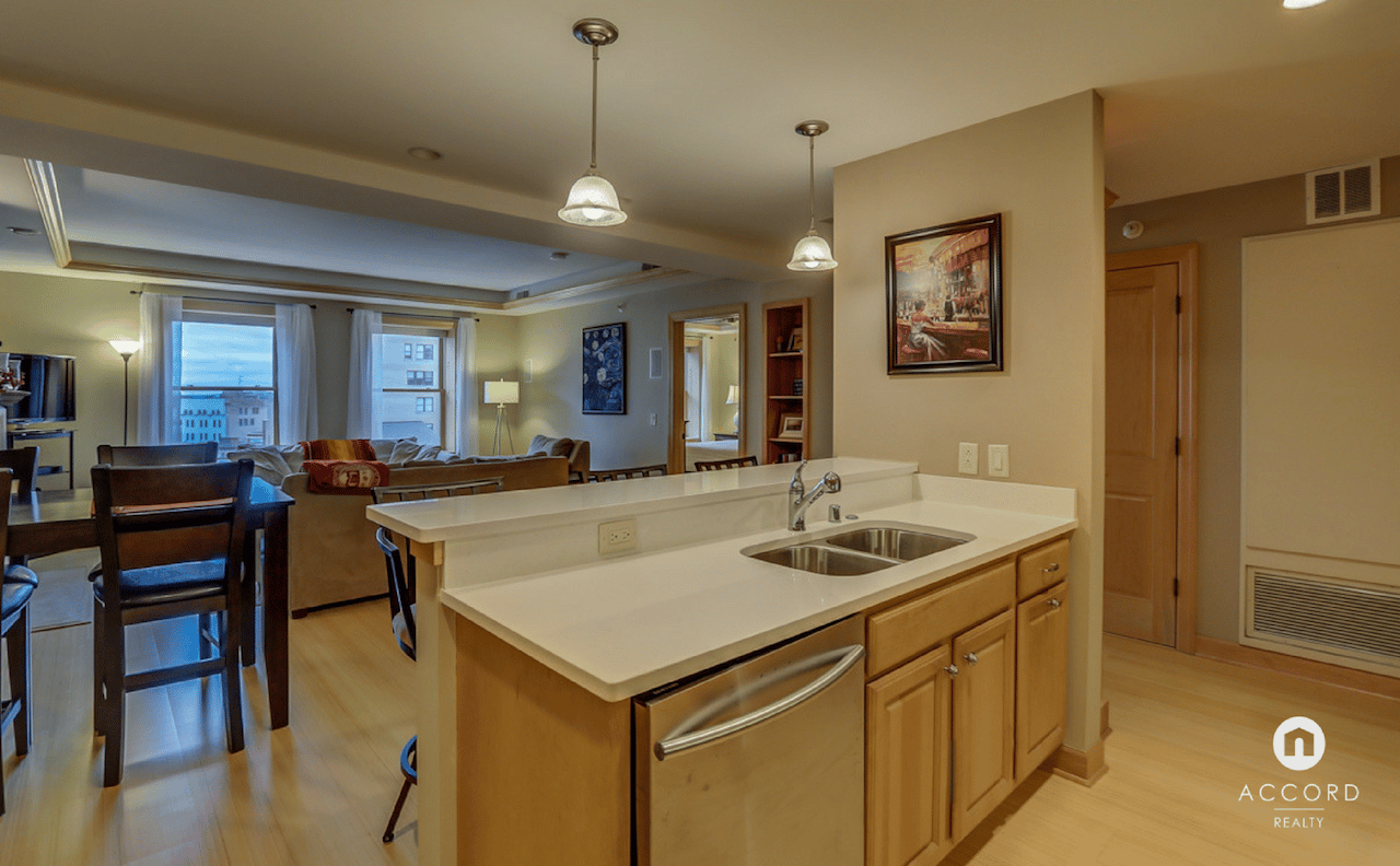 Open-plan condo kitchen with white peninsula, light wood cabinets, and living room beside city-view windows.